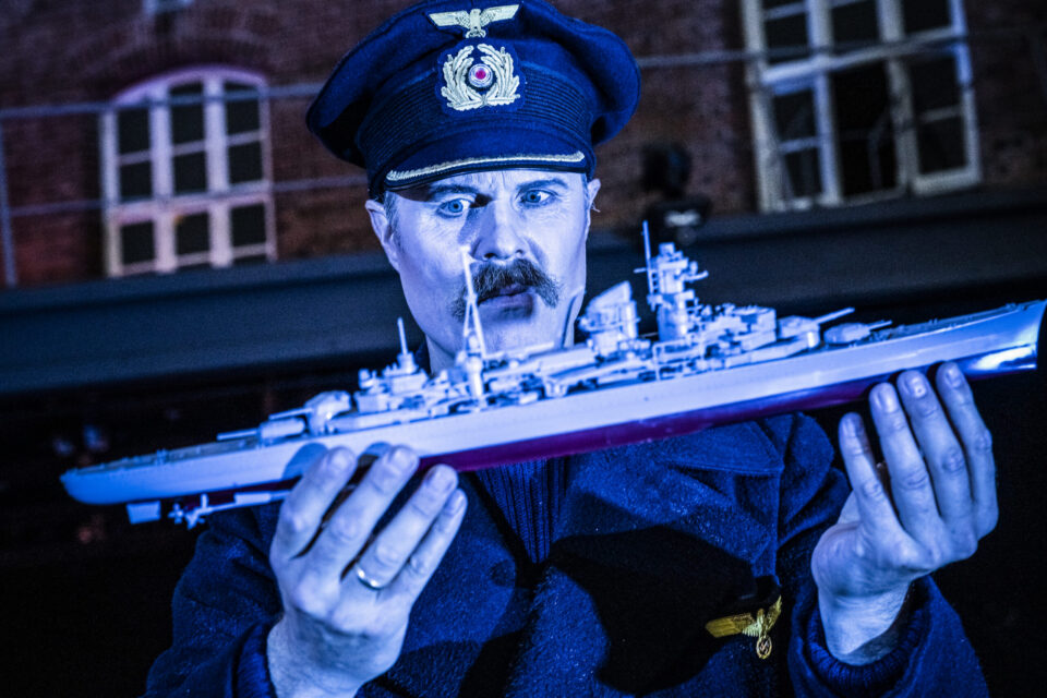 Hvit mann med skarp militæruniform som minner om 1940-tallets tyske uniformer holder et miniatyrskip i hendene. Stirrer på det. Bildet er preget av blålig lys. Skipet ser ut som en miniatyrutgave av den tyske krysseren Blücher, som ble sunket i Oslofjorden i 1940.