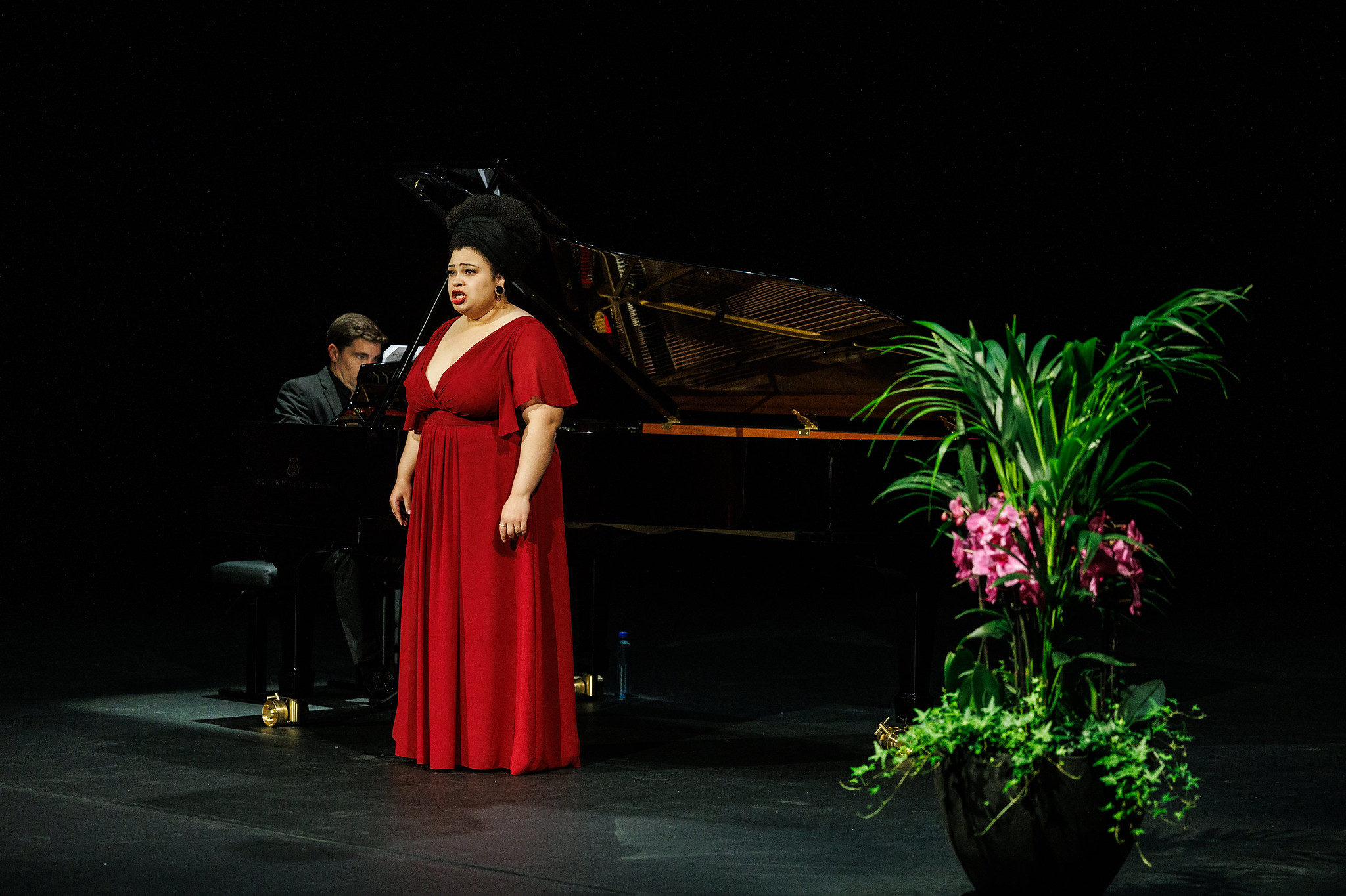Afrikansk-amerikansk kvinne i rød kjole synger opera ved et flygel. Mann i sort dress spiller på flygelet
