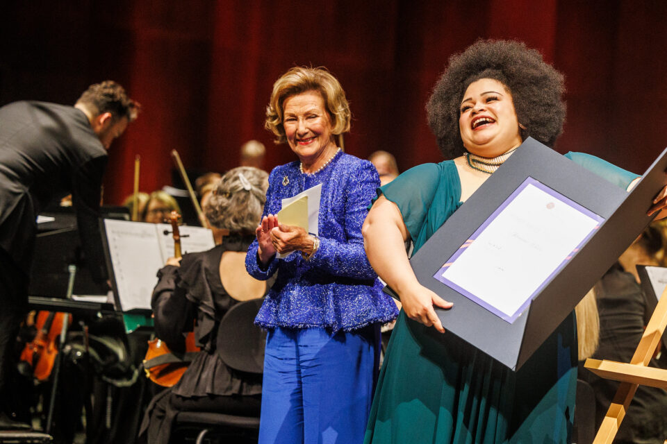 smilende dronning sonja i blå drakt, ved siden av afrikansk-amerikansk ung kvinne med diplom i hendene, smiler, begge står foran et orkester