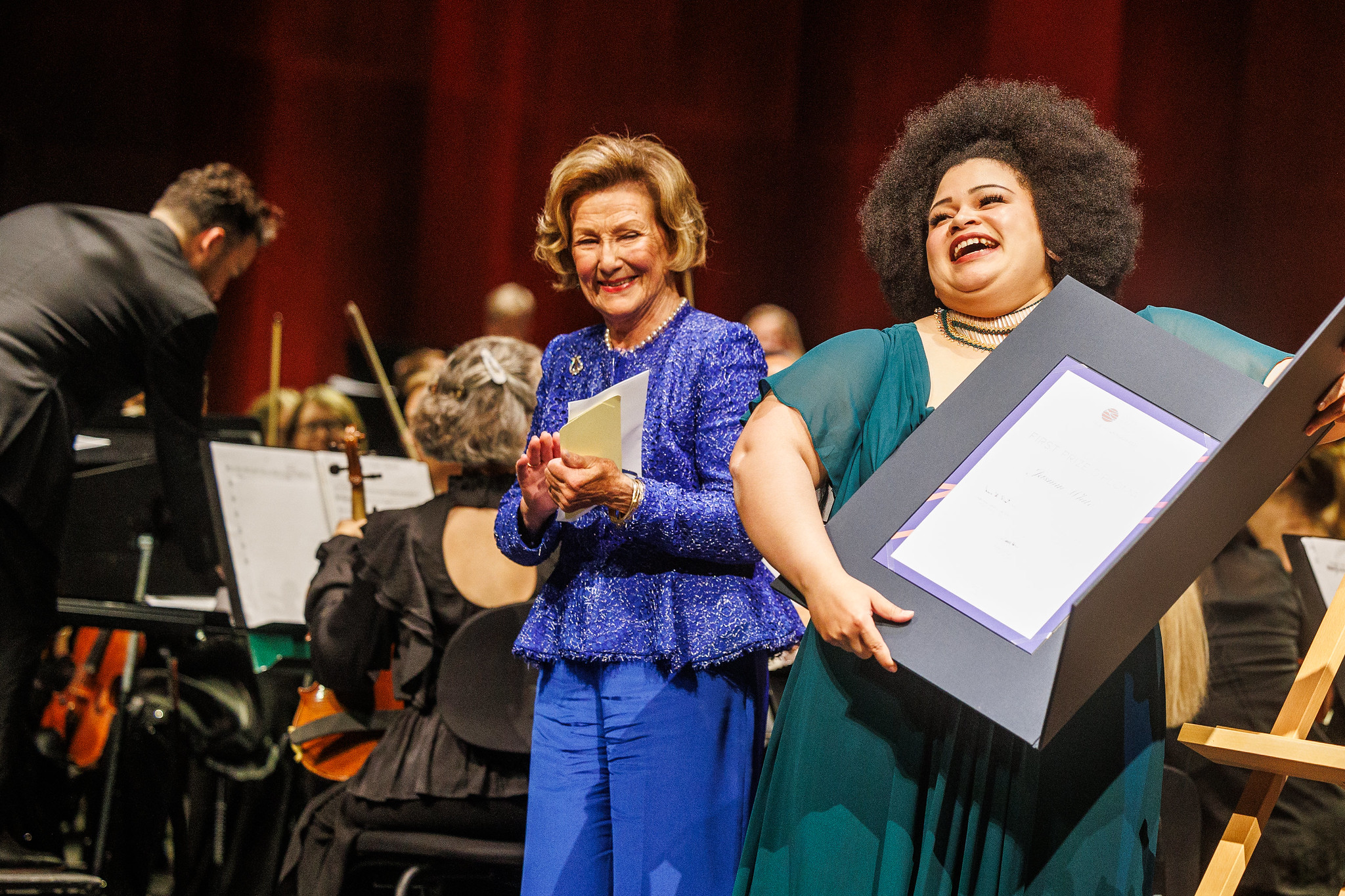 smilende dronning sonja i blå drakt, ved siden av afrikansk-amerikansk ung kvinne med diplom i hendene, smiler, begge står foran et orkester