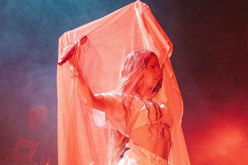 Flamencodanser og sanger under et slør som farges rødlig av scenelyset. Hun holder en mikrofon i venstre hånd.