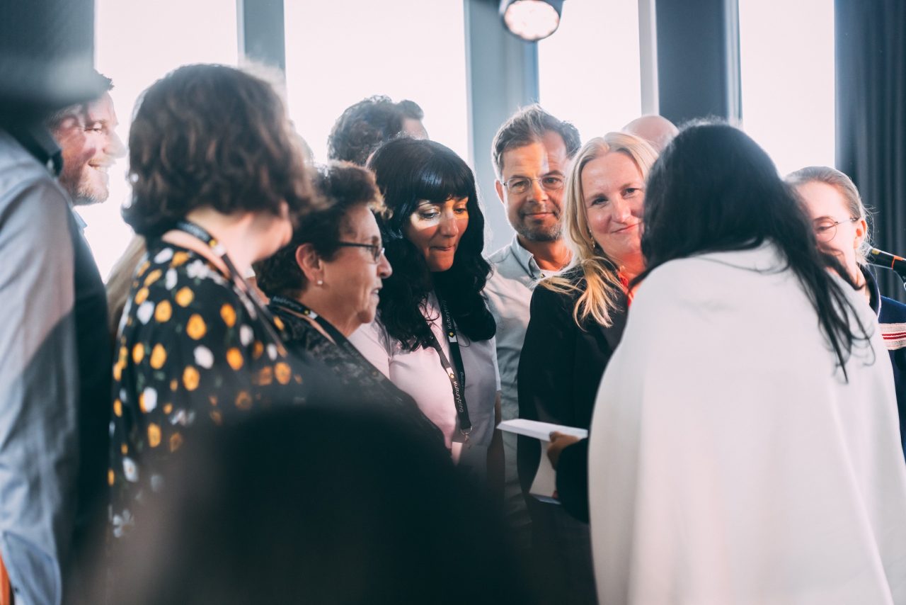 Professor i kultursosiologi Sigrid Røyseng (ser i kamera) gir rapporten til kulturminister Jaffery (med ryggen til). Ved siden av Røyseng, mot venstre, står utvalgsmedlem Sampda Sharma.